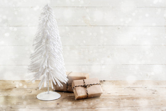 small christmas tree from white wire and gifts in kraft paper on a rustic wooden table, white painted vintage background with snowy bokeh lights, copy space