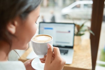 Rearview close up shot of a woman halding a cup of fresh coffee sitting at the cafe with her laptop...