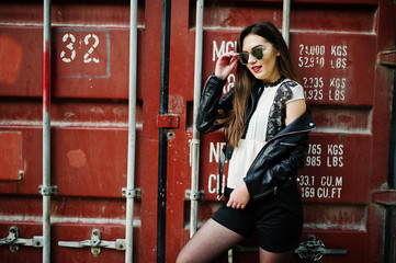 Stylish brunette girl wear on leather jacket and shorts, sunglasses against shipping container.