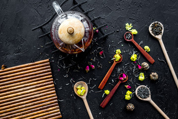 Brew the aromatic tea. Tea pot near wooden spoons with dried tea leaves, flowers and spices on black wooden background top view mockup