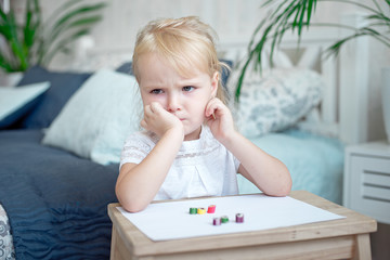 Sulky grumpy attractive little blond girl sitting with paper and crayons at a small wooden desk...