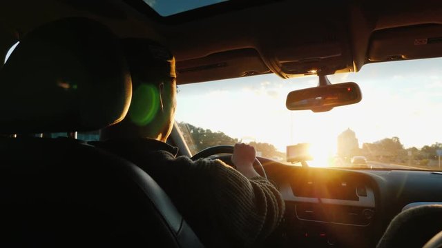 An Asian man drives a car along the highway, the setting sun shines in the windshield. Back view