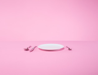 white plate, fork, spoon, and knife on a pink table in an abstract studio setting