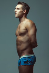 Fototapeta premium Male underwear fashion concept. Emotive portrait of young man in blue briefs posing with hands behind muscular body over blue background. Sportive style. Studio shot