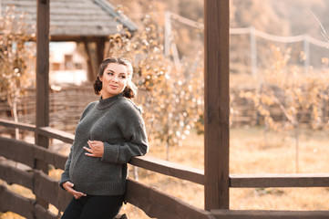 Happy pregnant woman wearing cozy knitted sweater leaning on wooden fence outdoors. Motherhood. Maternity. 20s.