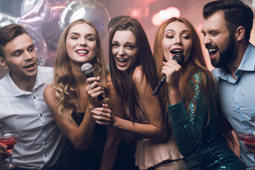 Three beautiful girls sing in a karaoke club. Behind them are men waiting for their turn. - obrazy, fototapety, plakaty