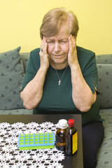 Starsza kobieta trzyma się za skronie z grymasem bólu na twarzy. Pojemniki z lekami oraz buteleczki leżą na stole obok niej. - obrazy, fototapety, plakaty