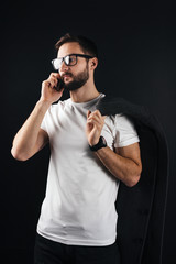 Young bearded hipster in glasses talking on smartphone standing in white blank thirt and keep coat on black background