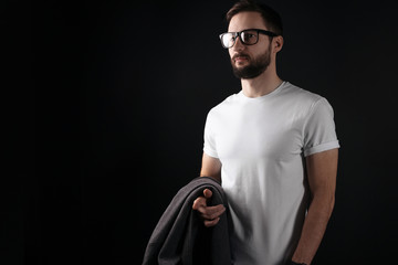 Beautiful bearded hipster wearing glasses and blank white tshirt and holds a coat in his hand on a black background