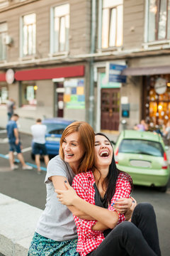 Two young girls hugging, laughting and sitting near road in the street. one brunette girl in red plaid shirt, another redhead girl wearing gray shirt and blue skirt. concept of sincere friendship
