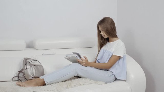 Cute girl sitting on sofa with tablet computer doing homework and playing games