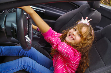 Close up of little beatiful smiling girl sitting in the car pretending to drive the luxury black car, weaing a pink blouse and jeans, in a blurred nature banground