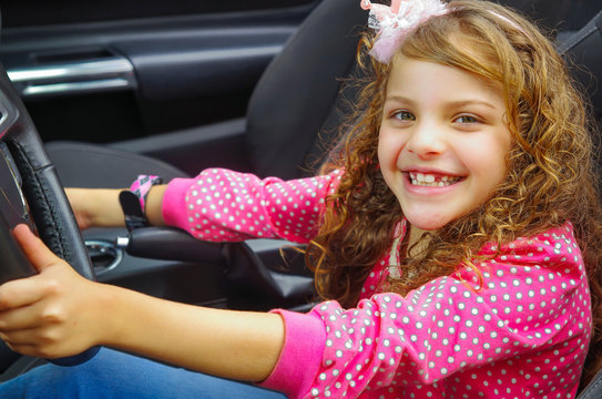 Close up of little beatiful smiling girl sitting in the car pretending to drive the luxury black car, weaing a pink blouse and jeans, in a blurred nature banground