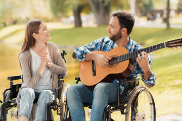Fototapeta na wymiar A man and a woman in wheelchairs met in the park. A man is playing the guitar, the woman listens and smiles.