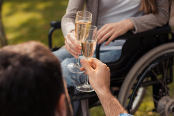 Close up. Woman in a wheelchair in the park with a man. A man is sitting on blanket. They clink glasses with champagne.