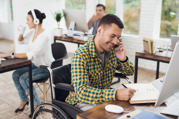 A man in a wheelchair writes with a pen in a notebook and talks on the phone. He is working in a bright office.