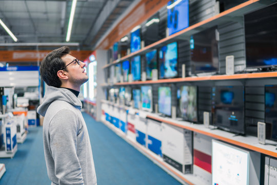 Smart modern male customer choosing large TV-sets at electronics store. He looks wondered. New screen generations.