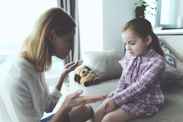 A woman punishes a girl and grimaces her finger. A frightened girl is sitting on the couch.
