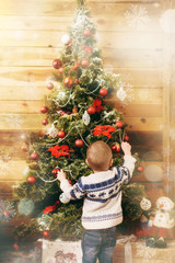 Happy little baby boy dressed in sweater decorating Christmas tree with toys in wooden room at home. Child good mood. New Year. Family and holiday 2018 concept. Magical highlight bokeh effect