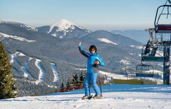 Full length shot of a smiling female skier showing thumbs up while skiing on the snowy slope at the winter ski resort. Mountains, forests, ski slopes and ski lifts on the background lifestyle concept
