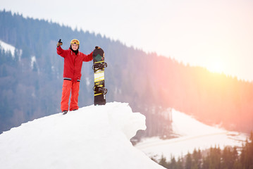 Man snowboarder standing on the top of the snowy slope with snowboard, smiling to the camera,...