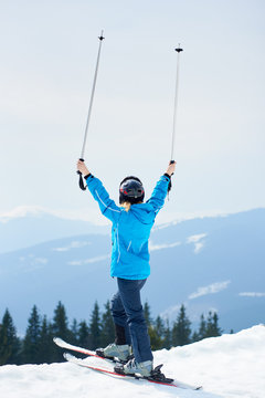 Back view of female skier wearing blue ski suit and black helmet, holding poles above a head, enjoying skiing at ski resort in the Carpathian mountains. Ski season and winter sports concept