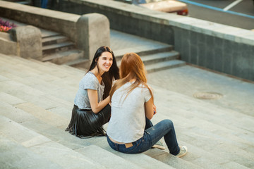 Fototapeta premium Two cheerful girls talking and sitting on the stairs in the street. one girl with long brunette hair in black skirt, another redhead girl in gray shirt and blue jeans. concept of sincere friendship
