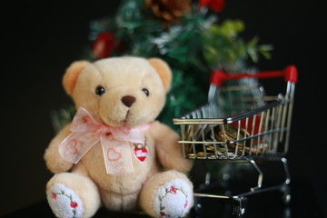 Cute fluffy teddy bear, coins in mini trolley and blurred Christmas tree isolated on black background. Saving, planning, shopping and celebration of Christmas day concept.