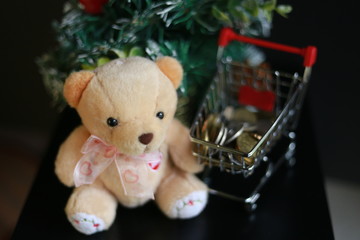 Cute fluffy teddy bear, coins in mini trolley and blurred Christmas tree isolated on black...