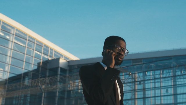 Attractive young African American businessman in a tie and glasses walking and talking on the mobile phone outside the big glass airport building on a sunny day