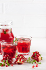Pomegranate cocktail with ice and mint in beautiful glasses and jug, fresh ripe pomegranate on white wooden background. Sweet red juice. Close up photography. Selective focus