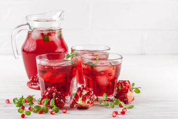 Pomegranate cocktail with ice and mint in beautiful glasses and jug, fresh ripe pomegranate on white wooden background. Sweet red juice. Close up photography. Selective focus