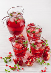 Pomegranate cocktail with ice and mint in beautiful glasses and jug, fresh ripe pomegranate on white wooden background. Sweet red juice. Close up photography. Selective focus