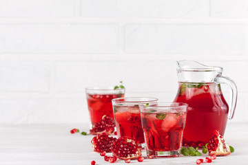 Pomegranate cocktail with ice and mint in beautiful glasses and jug, fresh ripe pomegranate on white wooden background. Sweet red juice. Close up photography. Selective focus