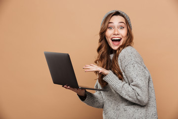 Happy exited attractive girl in warm clothes holding laptop computer while looking at camera