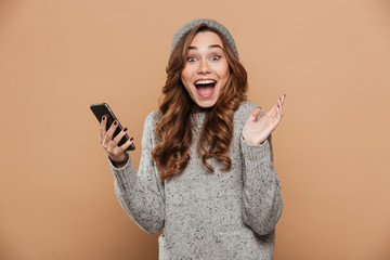 Portrait of happy exited brunette girl in gray hat holding mobile phone while looking at camera