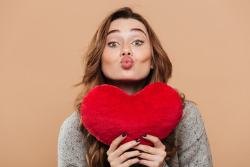 Close-up photo of lovely brunette girl holding big soft red heart while sending kiss, looking at...
