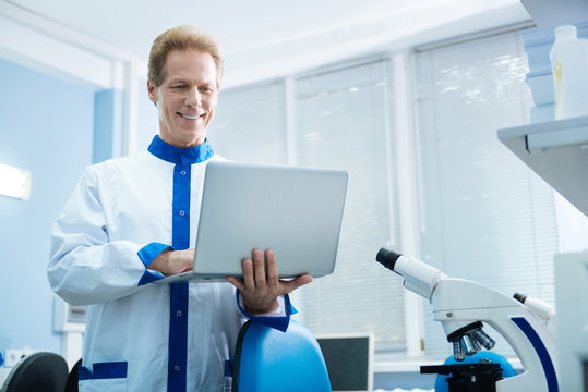 Conducting a research. Attractive inspired smiling biologist wearing a labcoat and holding a tablet while being in the lab and equipment in the background