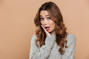 Young suprised brunette woman in gray pullover touching her cheek, looking at camera