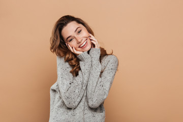 Portrait of cute smiling brunette girl in soft sweater touching her face while looking at camera