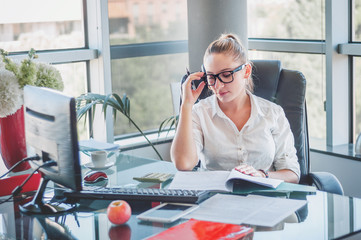 Portrait of young business lady in glasses sitting at her workplace and reads documents. Office worker at modern office workplace. Business concept.