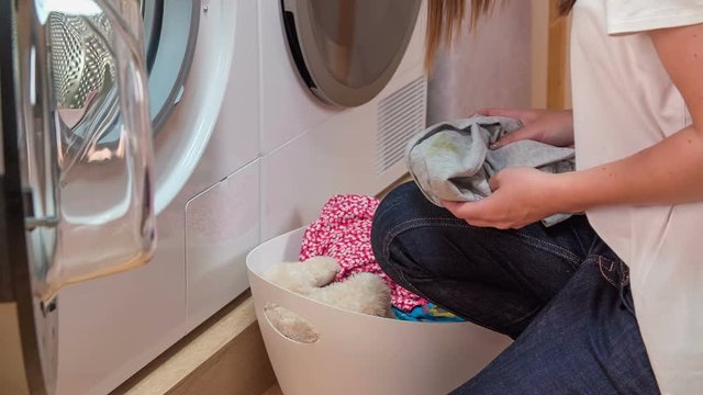Woman is gently and slowly putting laundry into washing machine. She is picking clothes from white basket.