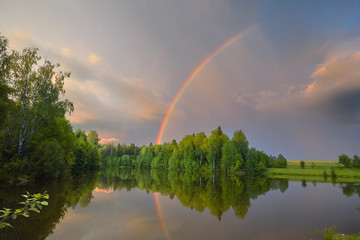 Rainbow over calm water of a forest lake. Beautiful clouds in the evening sky.