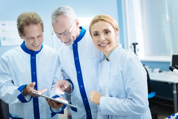 Team spirit. Smiling experienced scientists looking at the screen and wearing uniforms while content female blond researcher standing near them smiling