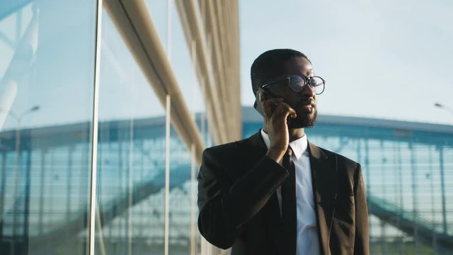 Close up of attractive young African American businessman in a suit and tie walking slowly along a modern glass office building and talking on the mobile phone. Office worker heading to his workplace