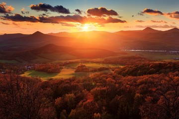 View from Plesivec hill, Ceske Stredohori, Czech. Beautiful sunset landscape with sun star. Evening villages. Sun with ping and orange sky. Green meadow, orange autumn trees in back light. Dark clouds
