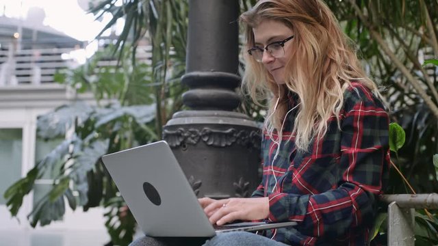 Cute and pretty young blonde woman in nerdy glasses sits in park with laptop, chats with friends, colleagues, listens to music from internet website through earphones, types messages to stay connected