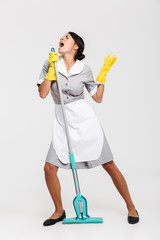 Full length portrait of funny housekeeper in uniform singing in mop as microphone