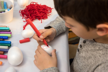 Young boy drawing heart on Styrofoam egg surrounded with crufts and arts material, sitting at table 