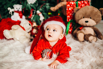 Fototapeta na wymiar Pretty little boy in red suit lies on fluffy carpet before a Christmas tree